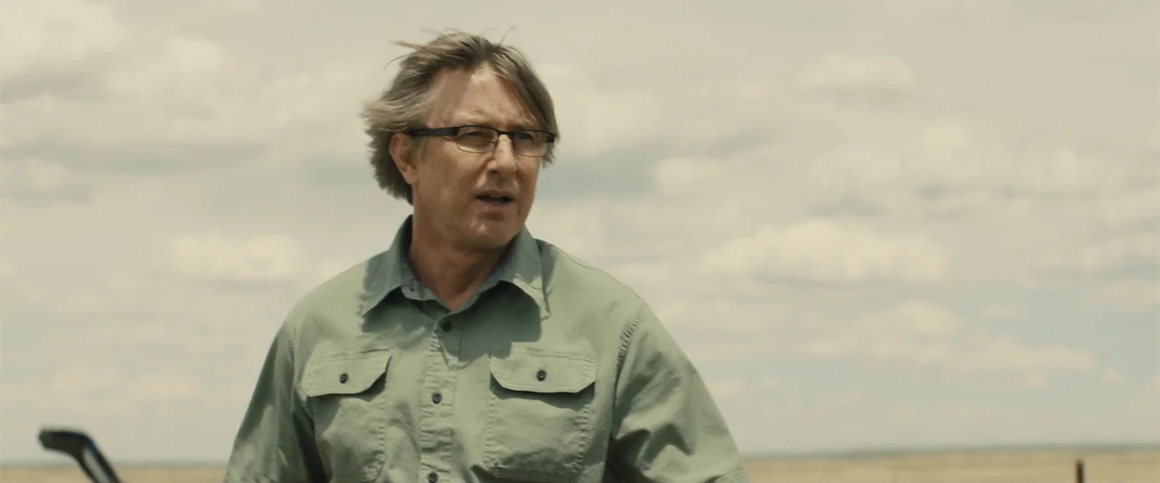 Those Who Wish Me Dead (2021) movie still: medium — An open rural field under an overcast sky provides a flat backdrop. A middle-aged man in a green…