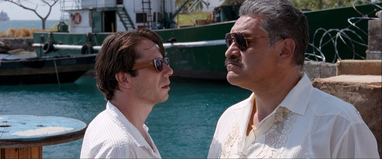 Quantum of Solace (2008) movie still: medium — A harbor scene with a large green boat docked against blue water. Two men stand in the foreground…