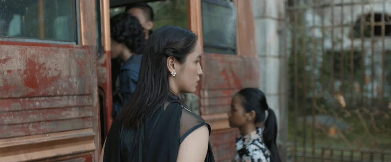 The Chanting 3 (2008) movie still: medium — A weathered red bus stands parked near a rusted metal fence in an outdoor space. A woman in a black top…