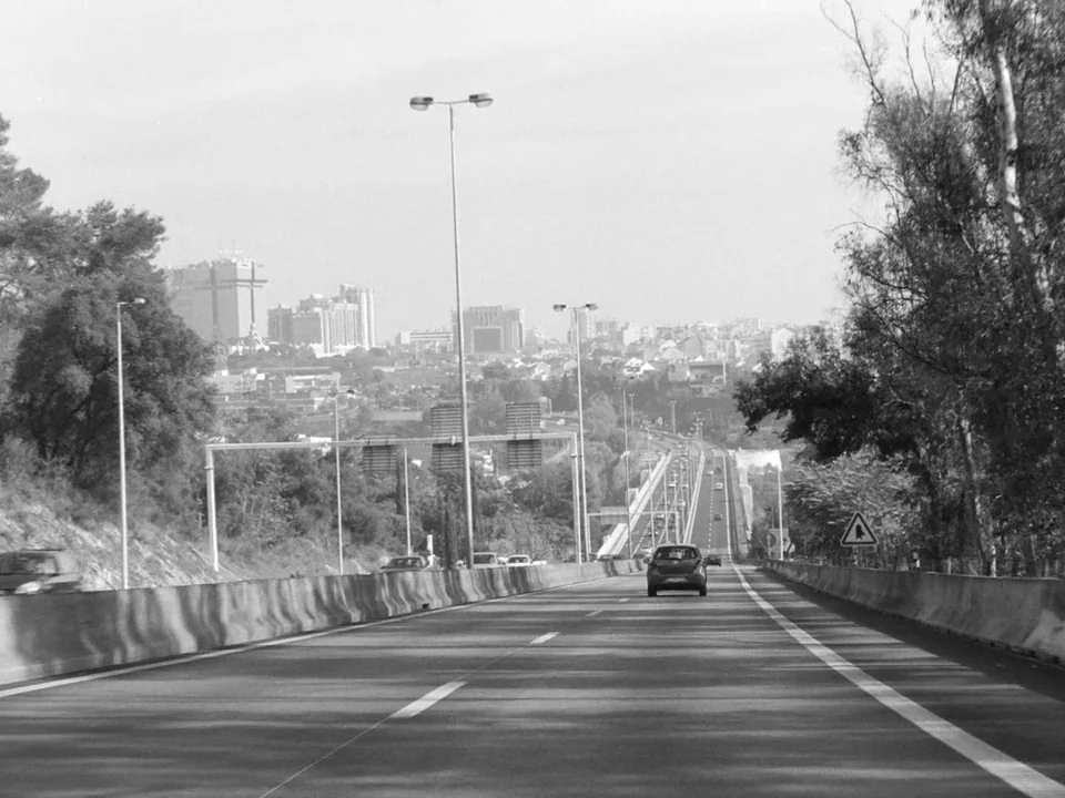 Tabu (2012) movie still: establishing — A paved multi-lane highway extends toward a city skyline under a clear sky. Concrete barriers line the road…