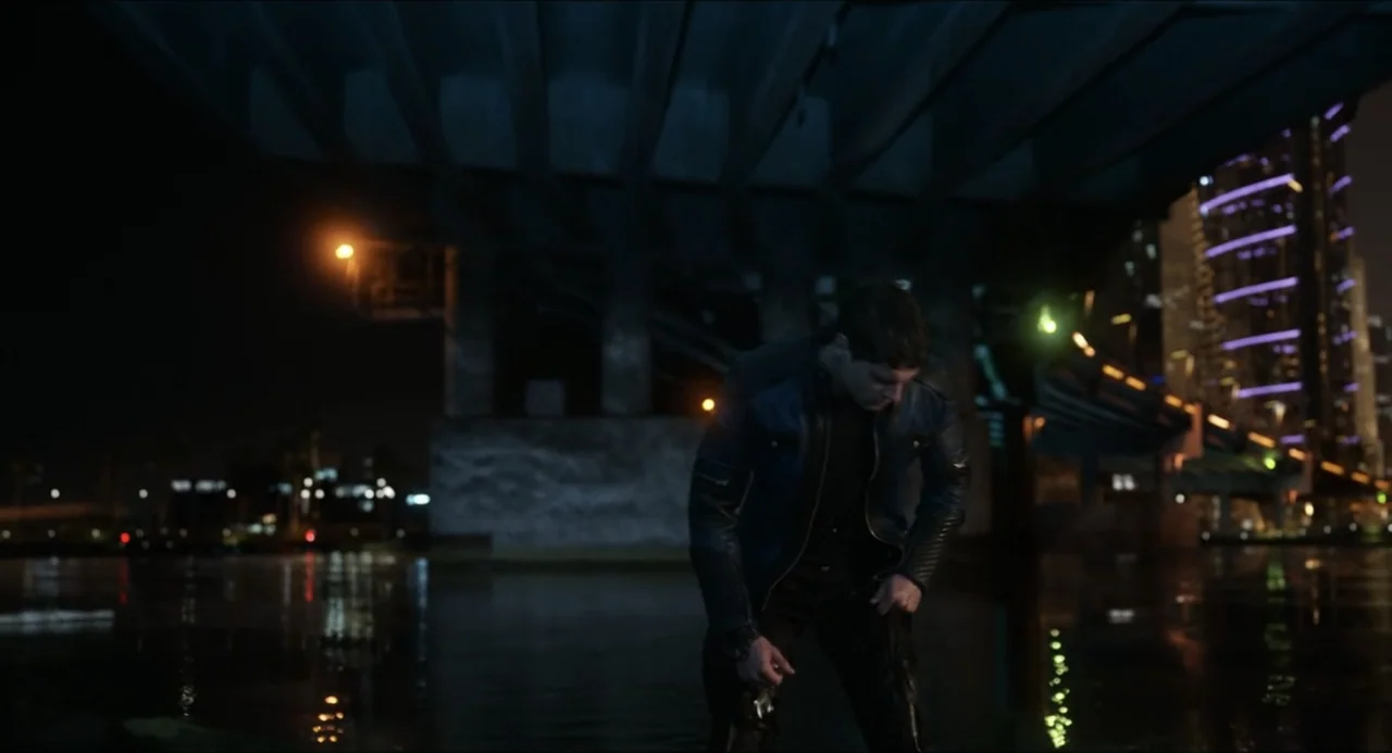 Henry Danger: The Movie (2025) movie still: medium — A dark riverbank area under a concrete overpass at night with city lights visible. A man in a…