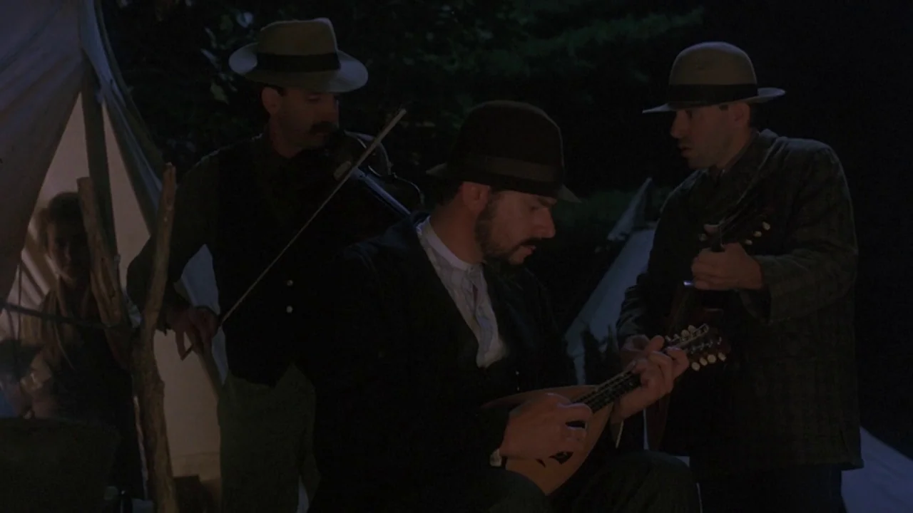 Matewan (1987) movie still: medium — Three men play instruments at a dark encampment next to a fabric tent. The musicians perform folk music…