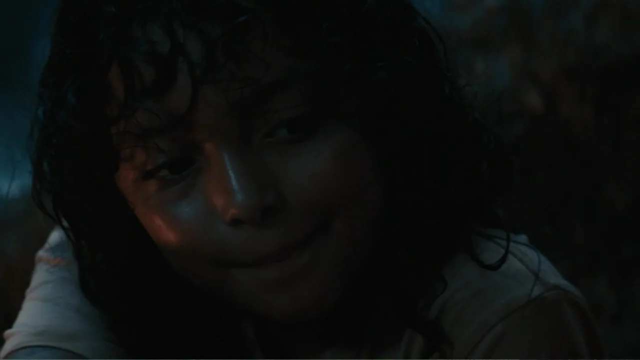Jurassic World Rebirth (2025) movie still: closeup — A dimly lit outdoor space at night with deep shadows obscuring the surroundings. A young boy…