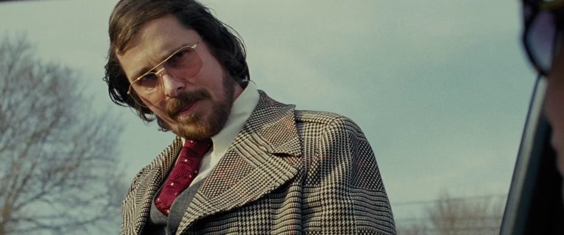 American Hustle (2013) movie still: medium — An outdoor parking lot area under an overcast sky with bare trees visible. A man in a houndstooth coat…