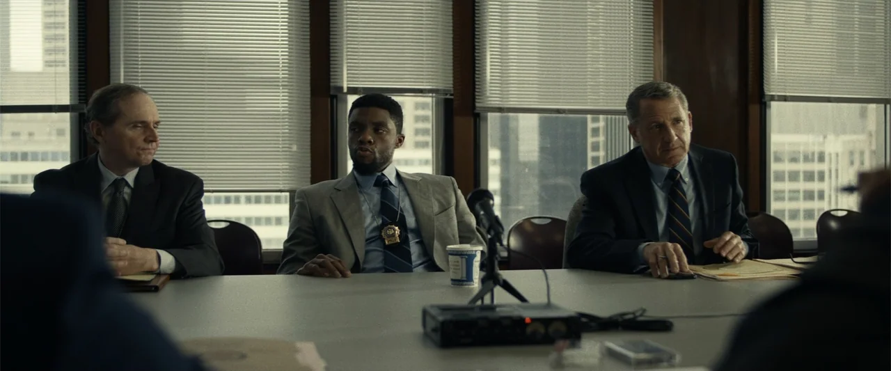 21 Bridges (2019) movie still: medium — A window-lined conference room features vertical blinds and a large table with audio equipment. Three men in…