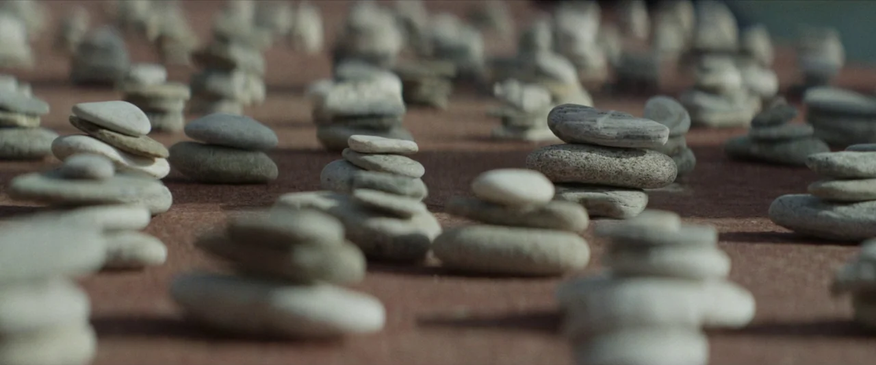 Chevalier (2015) movie still: closeup — Numerous small piles of stacked smooth stones rest on a reddish-brown textured surface. The depth of field…