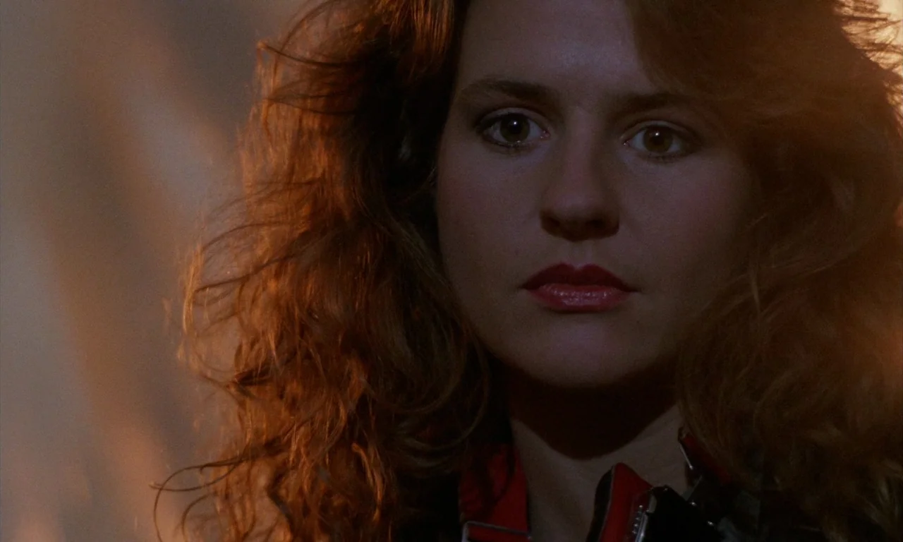 The Church (1989) movie still: closeup — A close-up shot of a young woman with voluminous hair in a dark space. The subject gazes ahead while bathed…