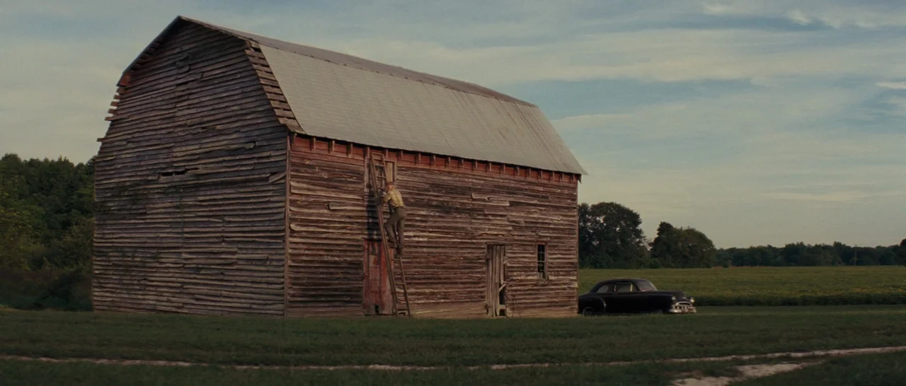 Loving (2016) movie still: wide — An aged, horizontal-plank barn stands in the middle of a grassy, open rural field. A lone man climbs a ladder on…