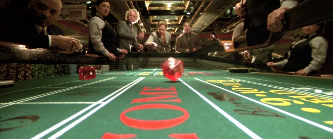 The Cooler (2003) movie still: wide — A green felt table surface dominates the foreground with red dice mid-toss. Several people in suits and…