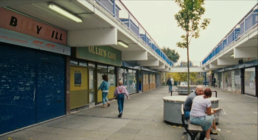 This Is England (2006) movie still: wide — An outdoor concrete shopping arcade features closed retail storefronts with graffiti and a cafe. Several…