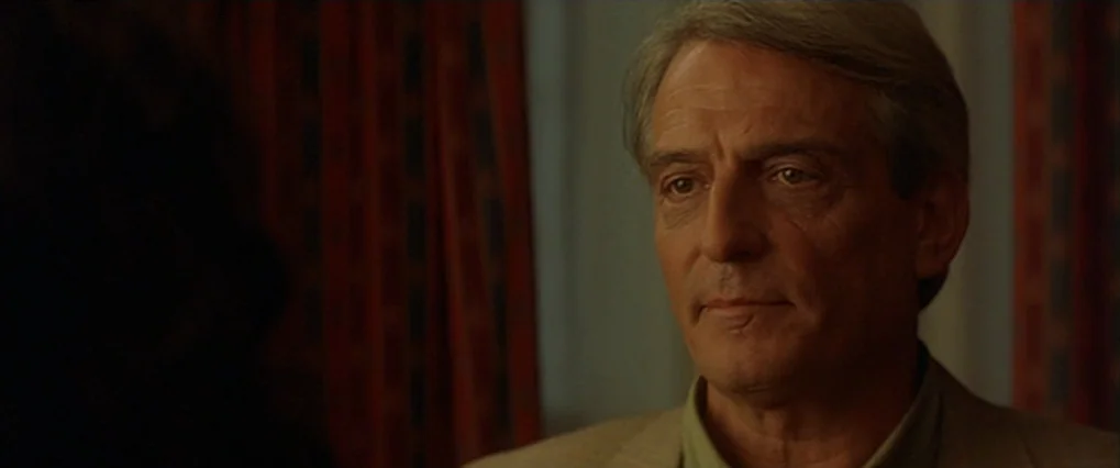Talk To Her (2002) movie still: closeup — A dimly lit interior space features dark red patterned curtains in the background. An older man in a light…
