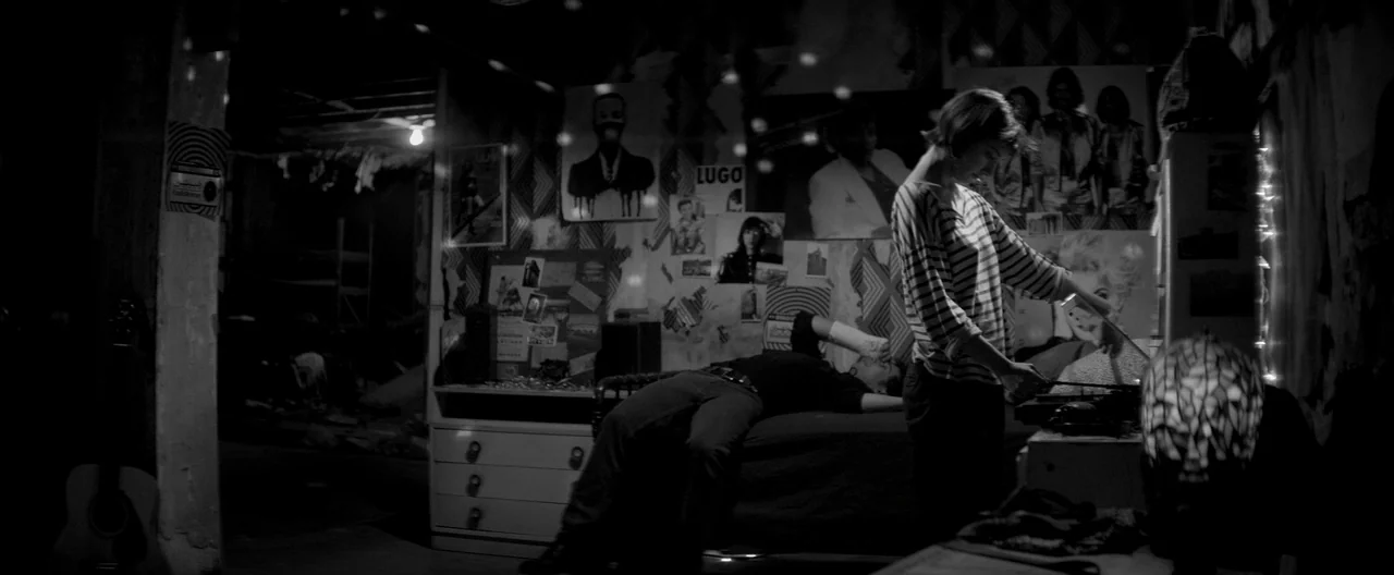 A Girl Walks Home Alone at Night (2014) movie still: medium — A cluttered bedroom is decorated with multiple posters on the walls. A woman interacts…