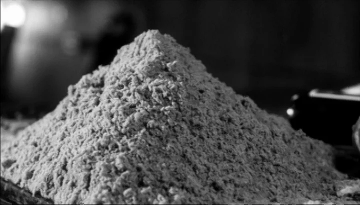 The American Astronaut (2001) movie still: closeup — A dense, conical mound of granular material rests on a dark, flat surface. The texture appears…