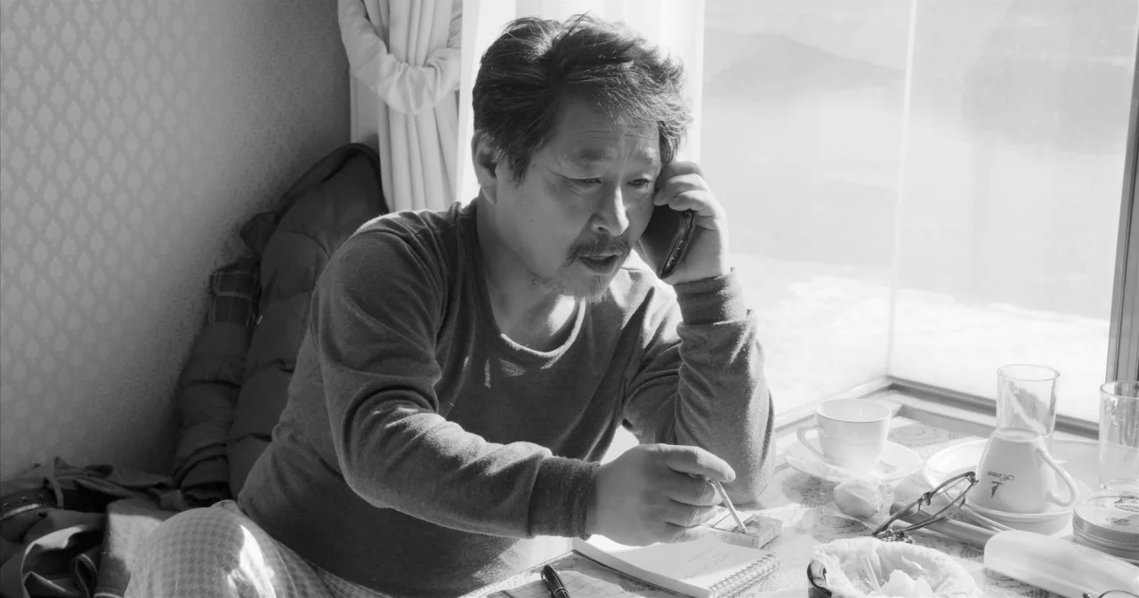 Hotel By The River movie still: medium — A man sits at a table beside a bright window during the day. He is talking on a mobile phone while writing…