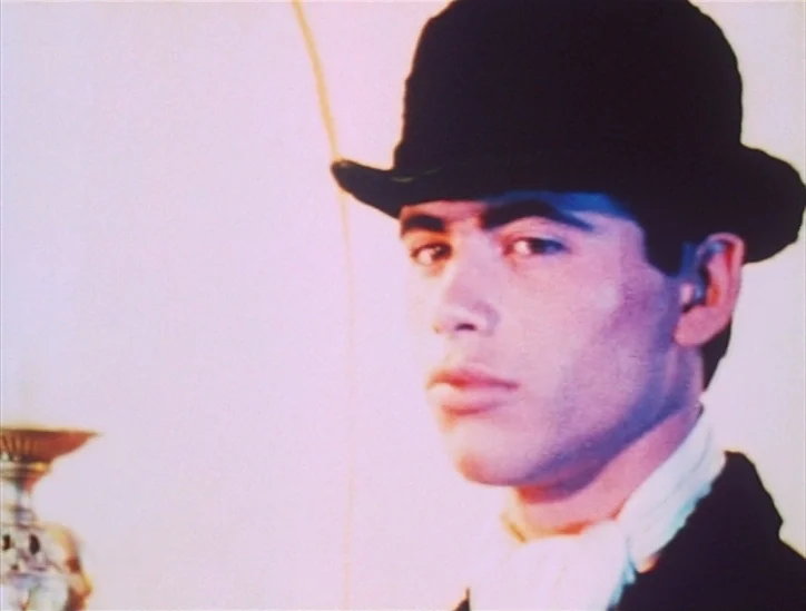 Pink Narcissus (1971) movie still: closeup — A man in a black bowler hat stands against a bright, featureless wall. He turns his head to gaze…