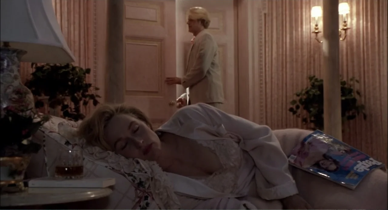 She-Devil (1989) movie still: medium — A dimly lit bedroom features floral wallpaper, sconce lighting, and a chaise lounge. A woman sleeps on the…