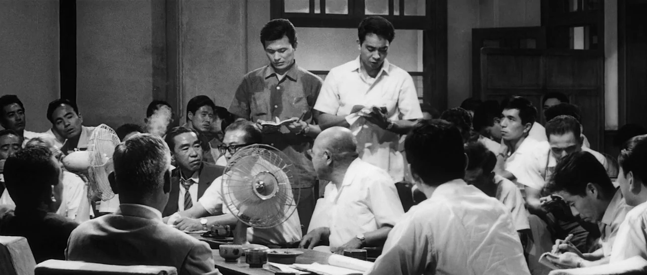 High and Low (1963) movie still: medium — A crowded, dimly lit office interior contains many men sitting around tables with fans. The men…