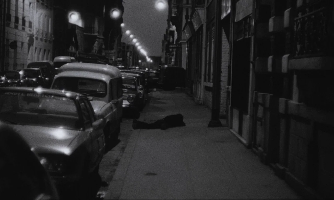 The Secret Son (1979) movie still: wide — A dark city street at night features a row of parked vintage cars. A lone human figure lies prone on the…