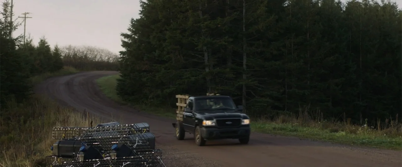 A Small Fortune (2021) movie still: wide — A black pickup truck travels down a narrow dirt road surrounded by dense pine trees. The vehicle carries…