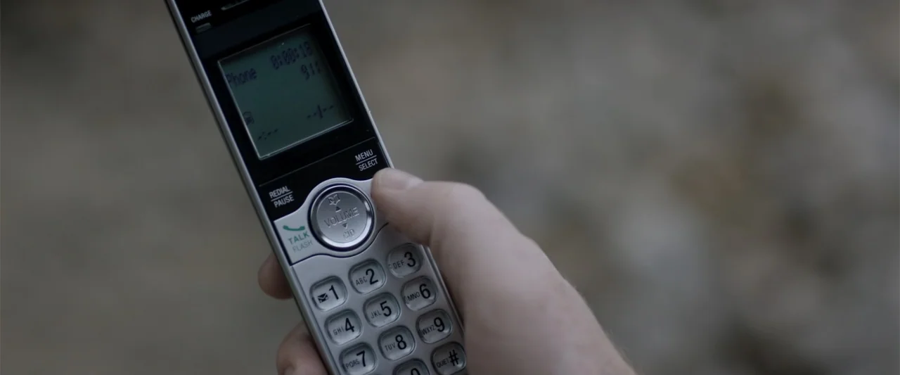 Radius (2017) movie still: closeup — A silver cordless phone is held in a person's hand against an out-of-focus background. The phone screen…