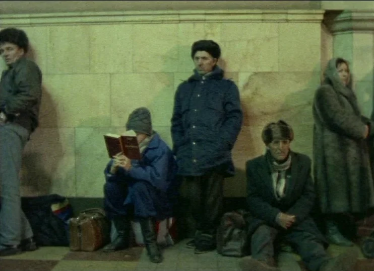 From The East (1993) movie still: medium — Commuters wait against a pale, tiled wall in a dimly lit, utilitarian public space. Several people are…