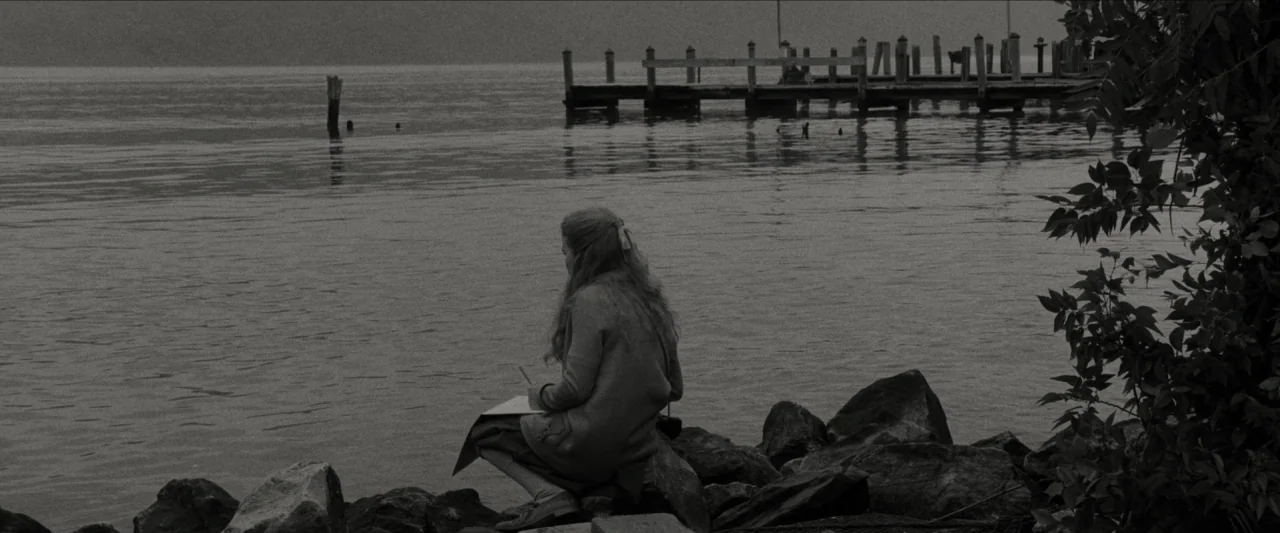 Wonderstruck (2017) movie still: wide — A calm, overcast lakefront features a wooden pier extending into the water. A young girl sits on large rocks…