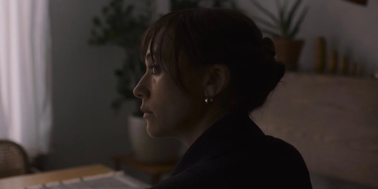 The Sound of Silence movie still: closeup — A dimly lit room contains a white curtain and several potted plants. A woman in a black top looks toward…