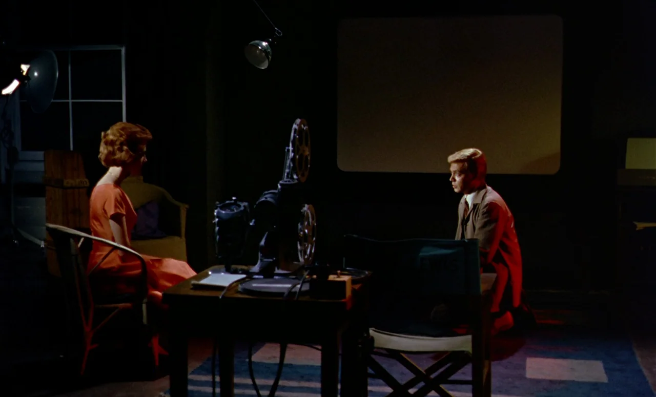 Peeping Tom (1960) movie still: wide — A dark, dimly lit room contains a central wooden table with a film projector. The woman in an orange dress…