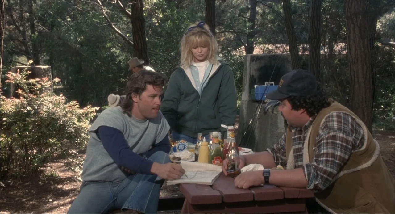 Overboard (1987) movie still: medium — A group gathers at a wooden picnic table located in a shaded wooded park. One man writes in an open notebook…