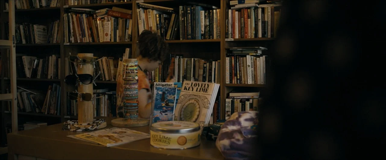 Road House (2024) movie still: medium — A cluttered shop interior features wall-to-wall shelves filled with books and various trinkets. A young…