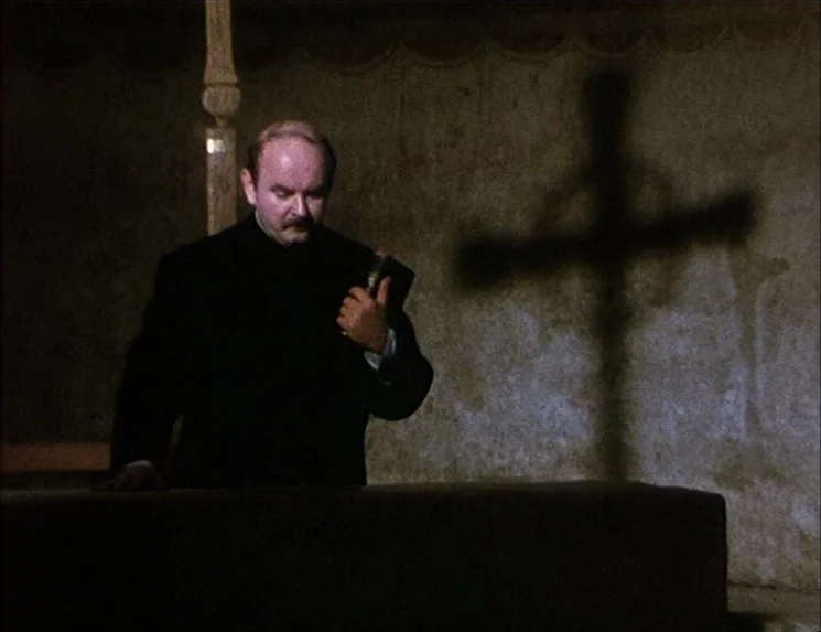 The Niklashausen Journey (1970) movie still: medium — A dim room features a textured wall with a sharp shadow of a cross. A man in a dark garment…