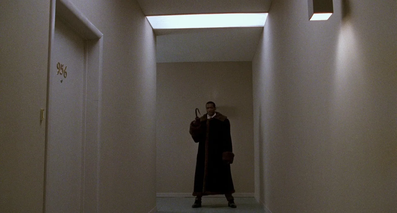 Candyman (1992) movie still: wide — A long, minimalist apartment building corridor features smooth beige walls and a numbered door. A man wearing a…