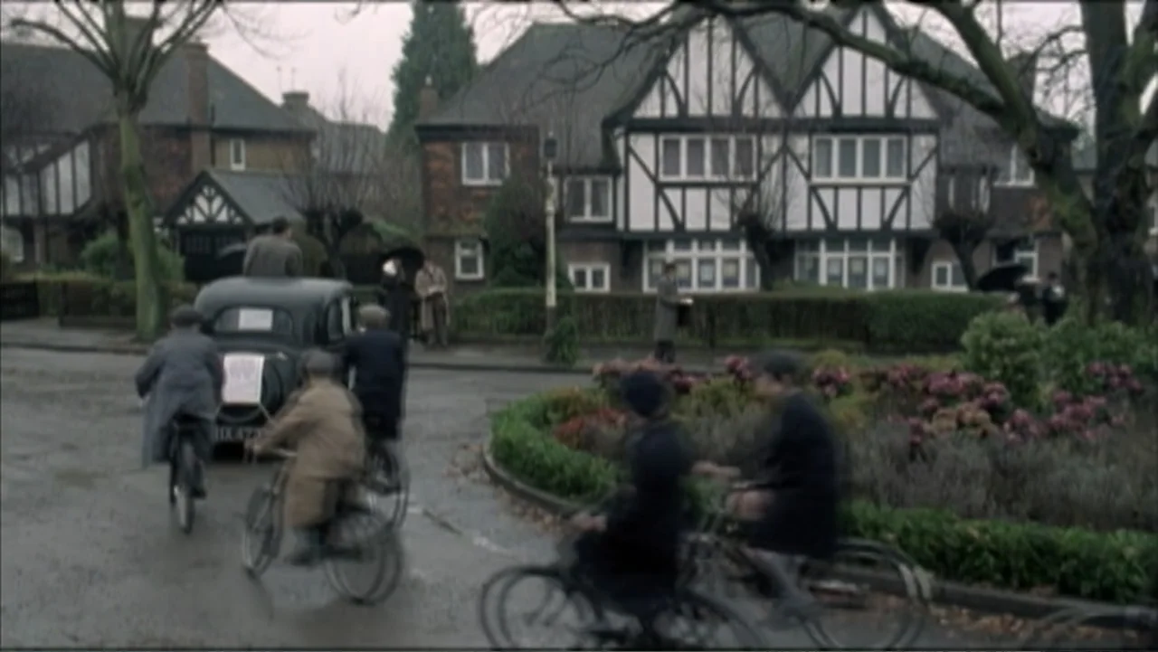 The Gathering Storm (2002) movie still: wide — A residential street lined with Tudor-style houses features a central circular planting bed. Several…