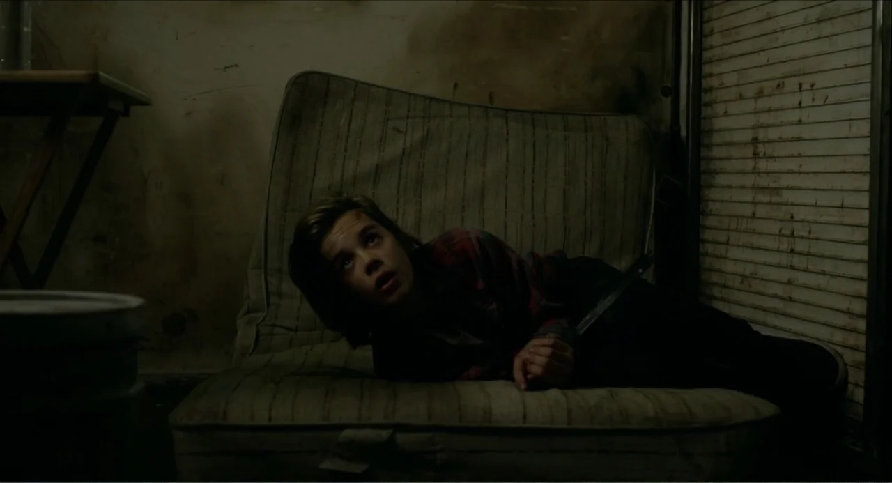Instantes Capturados (2022) movie still: medium — A dim room contains a stained, worn mattress on the floor near a metal shutter. A teenage boy with…