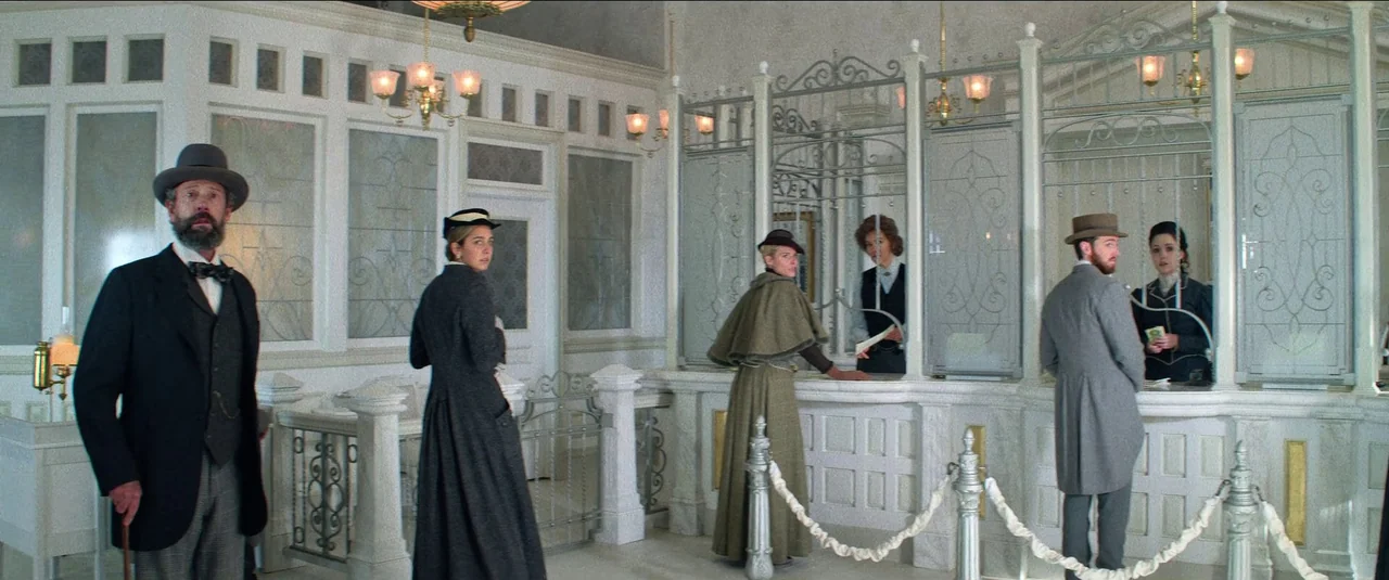 The Harder They Fall (2021) movie still: wide — A spacious 1910s era bank interior features ornate white marble counters and glass partitions. Four…
