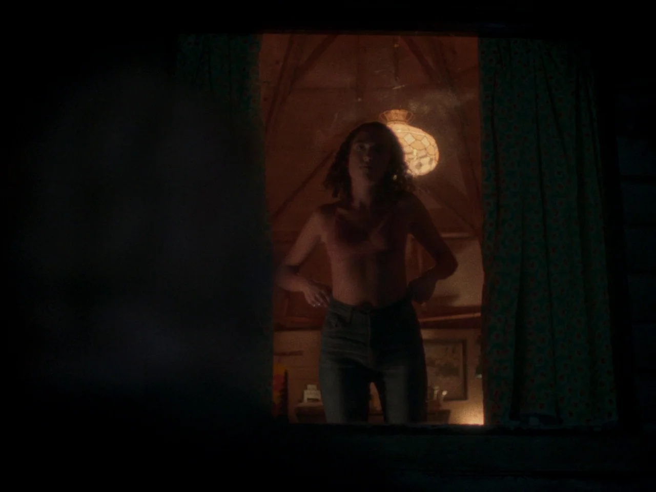 In A Violent Nature (2024) movie still: medium — A dimly lit room with floral curtains and a wooden ceiling structure. A young woman adjusts her…