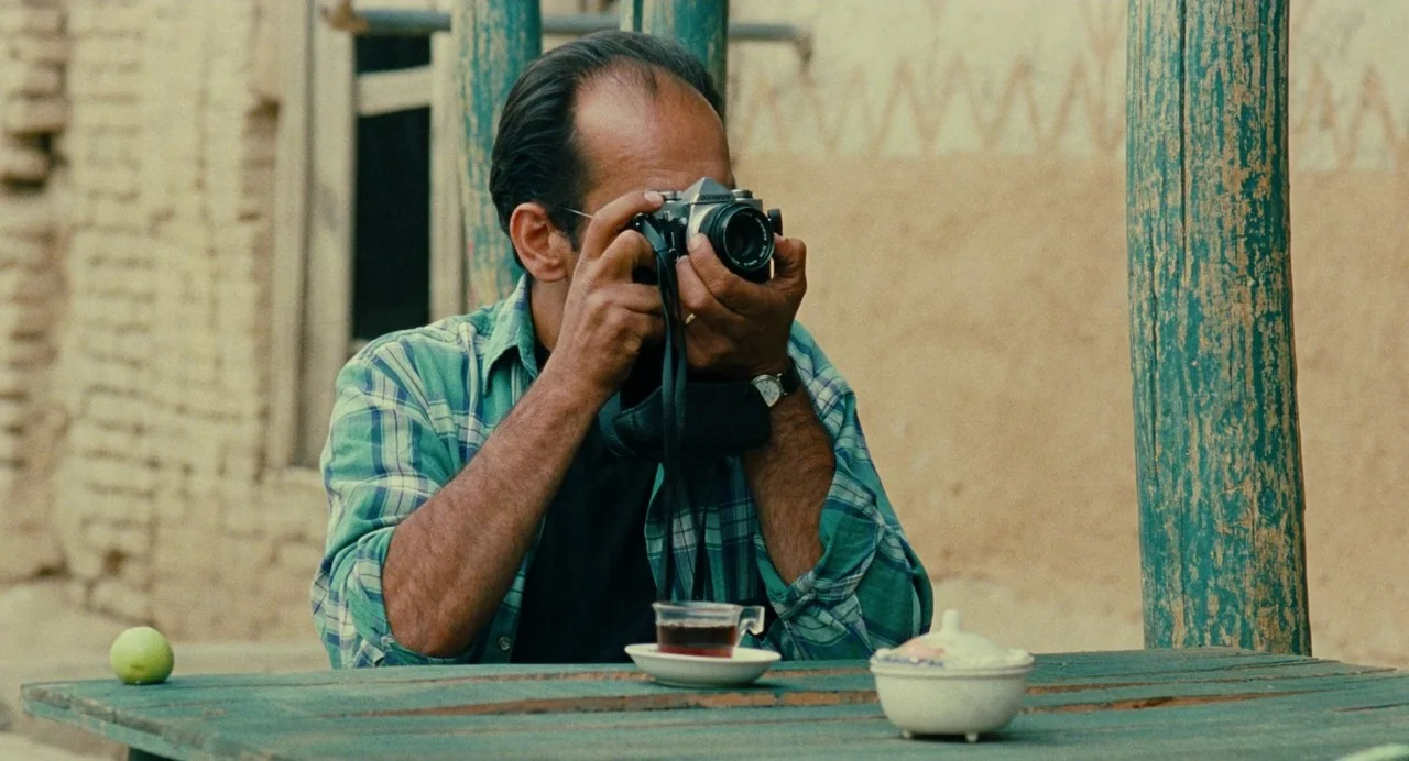 The Wind Will Carry Us (1999) movie still: medium — A man sits at a weathered green wooden table in front of a mud-brick wall. The man holds an…