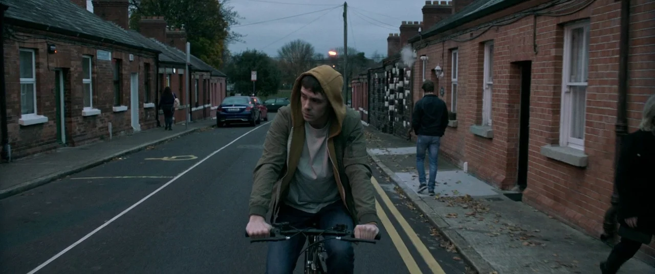 The Cured (2017) movie still: medium — A narrow road lined with identical brick row houses under an overcast sky. A young man in a tan hoodie rides…