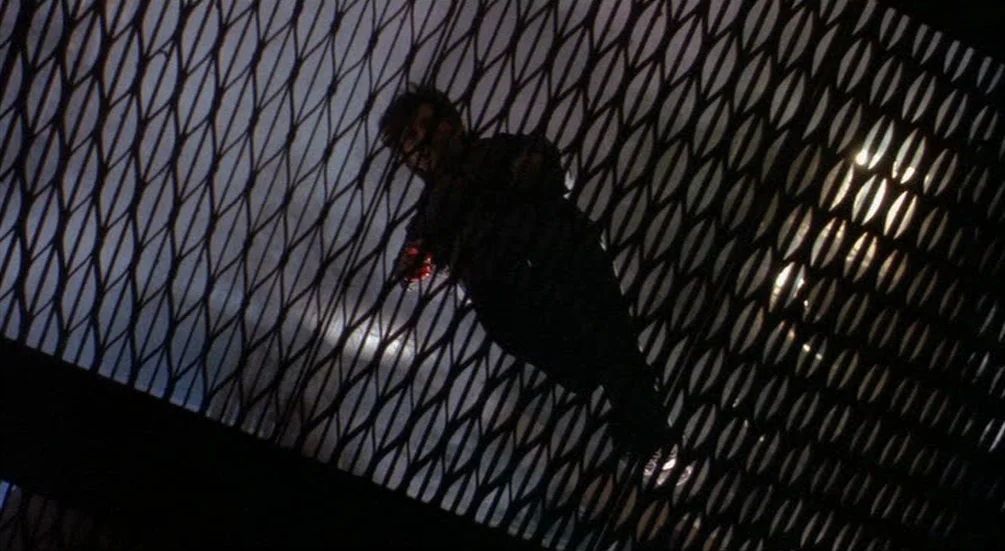 Freddy’s Dead: The Final Nightmare (1991) movie still: medium — A dark industrial metal mesh catwalk obscures a person standing in the background.…