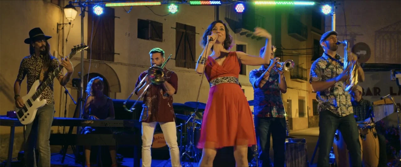 A Hipster in Rural Spain (2024) movie still: fullbody — A musical group performs on a makeshift stage set up in a town square at night. The members…