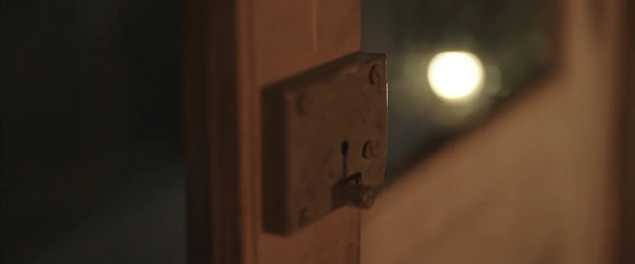 Peranbu (2019) movie still: closeup — A close-up view shows a vintage metal door lock mechanism mounted on a wood door. A glowing blurred light…