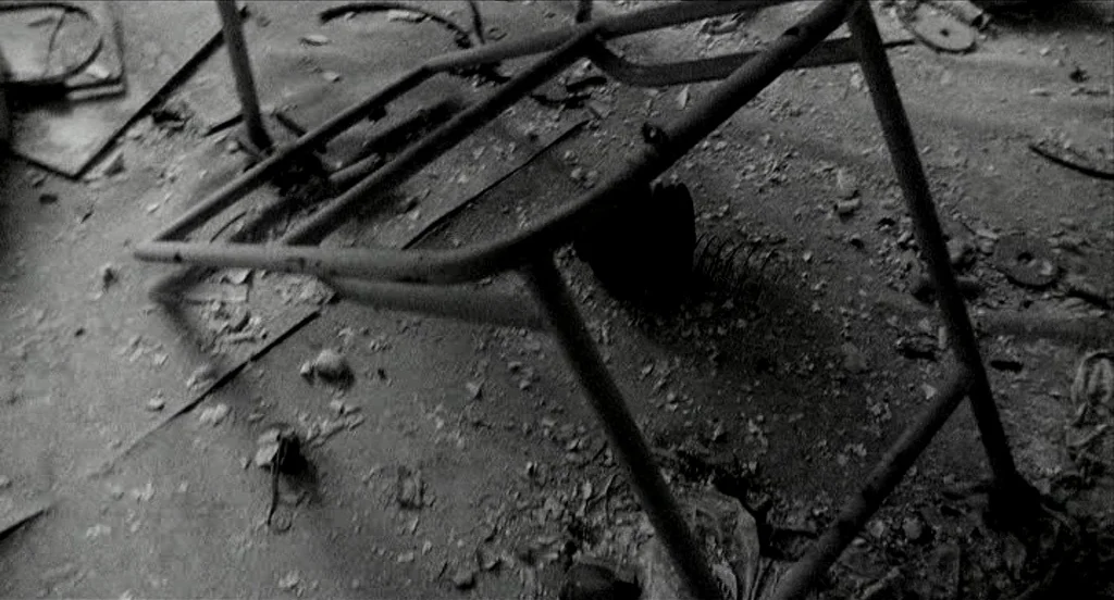 Container (2006) movie still: closeup — A metal frame rests on a floor covered in dusty construction debris and rubble. The scene is shot in…