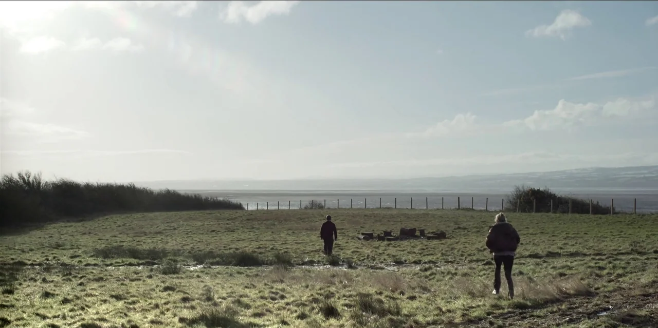 Help (2021) movie still: wide — A wide, grassy meadow stretches toward a coastal inlet lined by a simple wire fence. A man and a woman walk across…