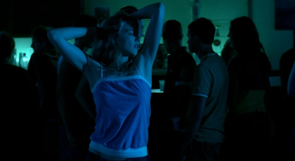 Water Lillies (2007) movie still: medium — A dimly lit indoor gathering with multiple silhouetted figures standing in the background. A young woman…