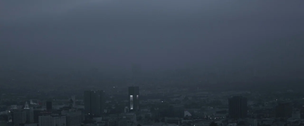 Starry Eyes (2014) movie still: establishing — A dense urban skyline is obscured by heavy low-hanging clouds and atmospheric haze. Several dark…