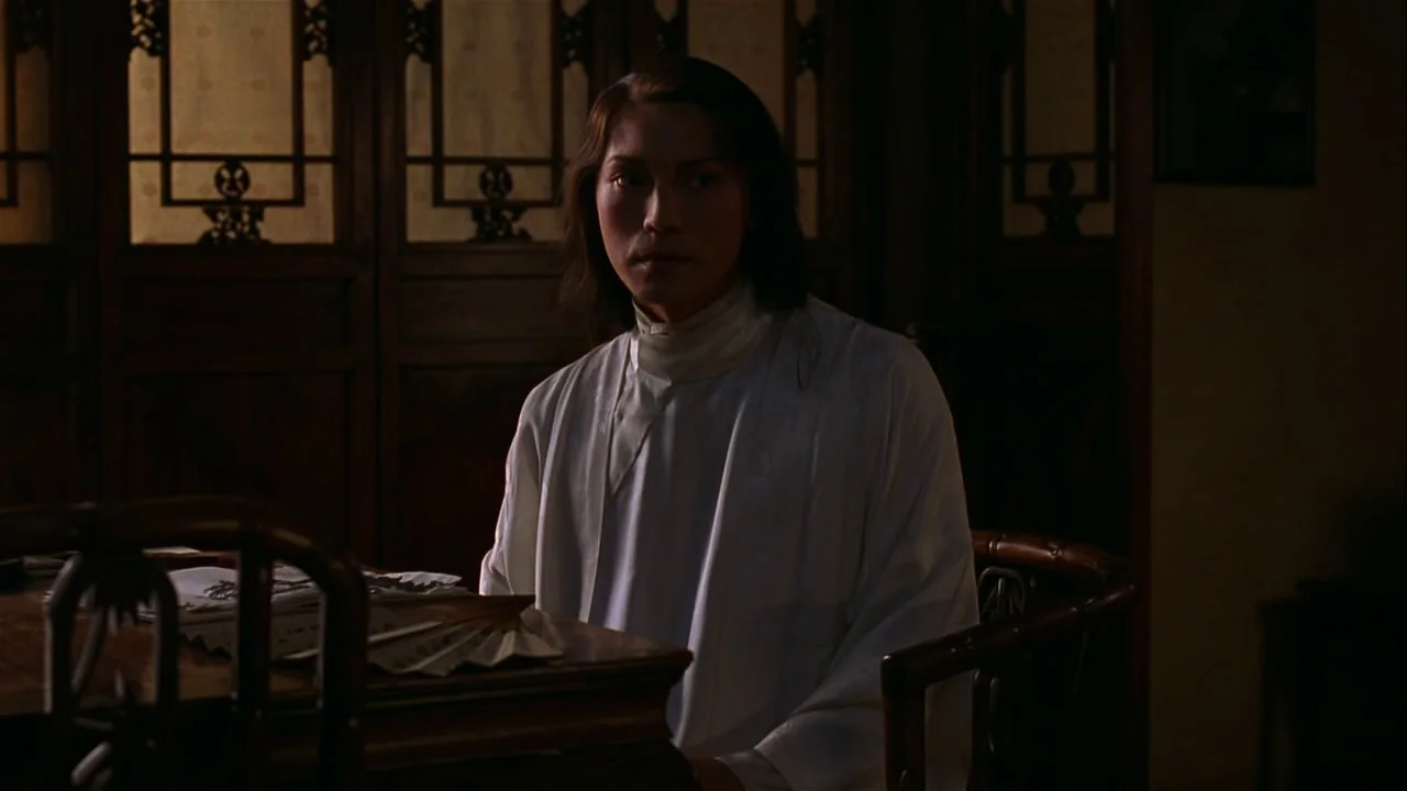 M. Butterfly (1993) movie still: medium — A dimly lit room features a wooden table and a traditional lattice screen background. A young man sits at…