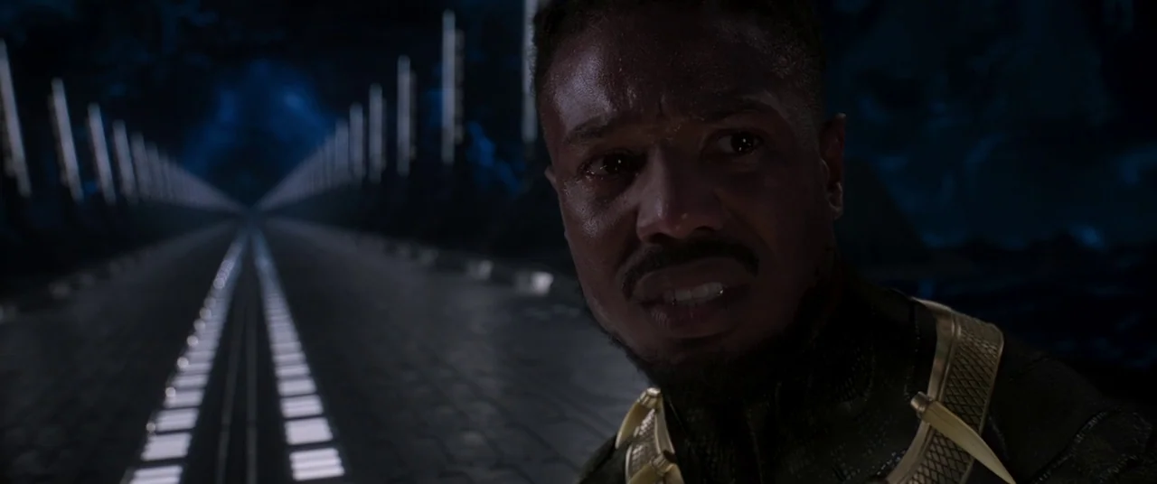 Black Panther (2018) movie still: closeup — A dark, long corridor stretches into the distance with blue floor lights. A man with a dark suit and…