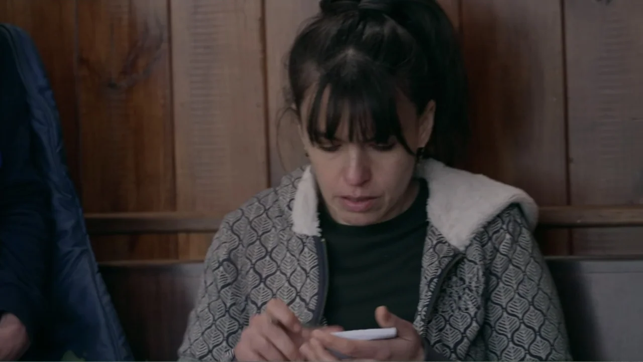 Saturdays Disorders (2022) movie still: medium — A woman sits in front of a wooden panelled wall. She uses a smartphone while looking downwards.