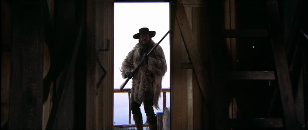 McCabe & Mrs.Miller (1971) movie still: fullbody — A dark wooden structure viewed from within looking out through an open doorway. A man stands in…