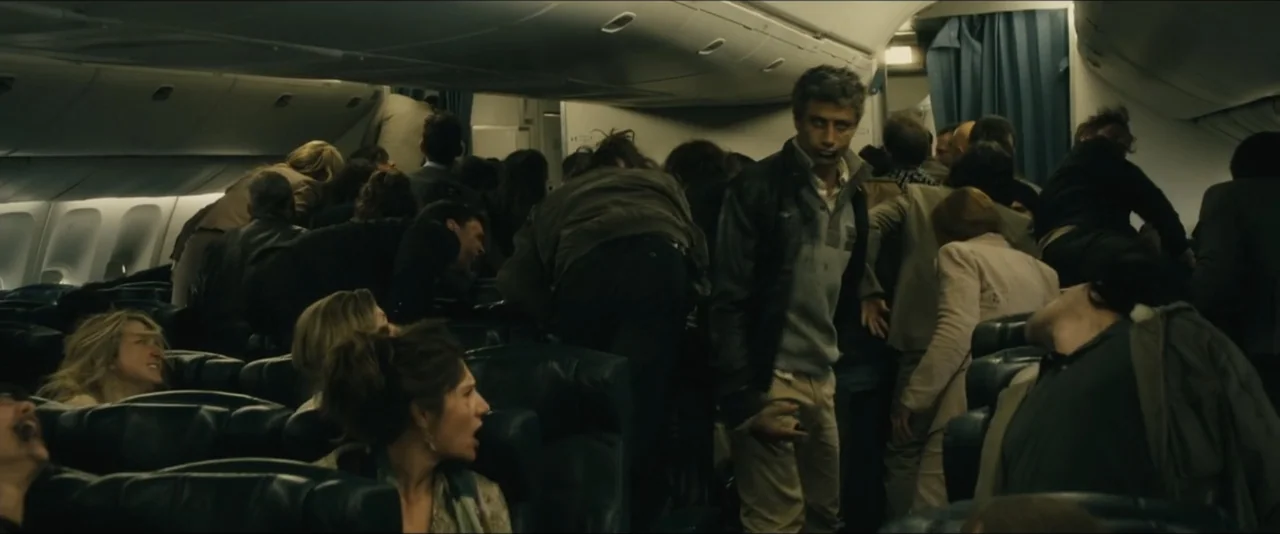 World War Z (2013) movie still: medium — The interior of a cramped commercial airplane cabin is filled with distressed, reacting passengers. A man…